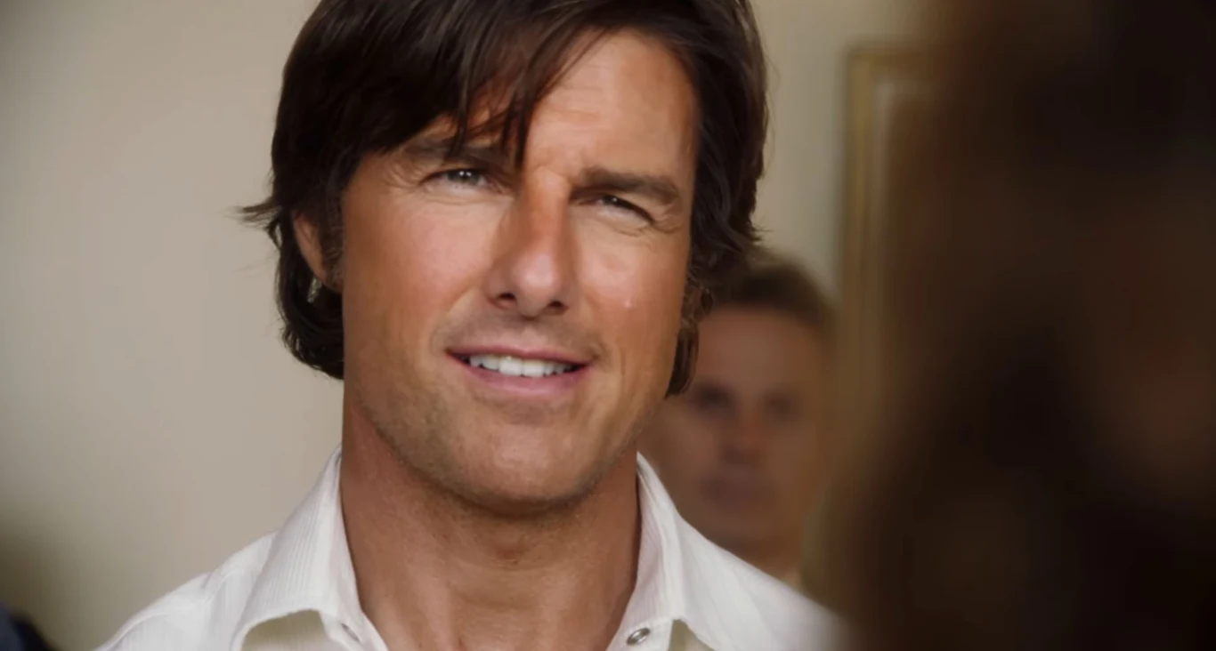 Tom Cruise w filmie "Barry Seal: Król przemytu" Mężczyzna w jasnej koszuli, z ciemnymi włosami, stoi we wnętrzu z rozmytą postacią w tle