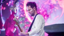 Mężczyzna w białej koszuli grający na gitarze basowej podczas występu scenicznego, w tle kolorowe oświetlenie i rozmyty, wielobarwny motyw świetlny.