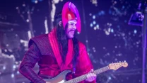 Gitarzysta w oryginalnym kostiumie sceniczny z czerwonymi elementami i hełmem o futurystycznym kształcie gra na elektrycznej gitarze podczas koncertu.