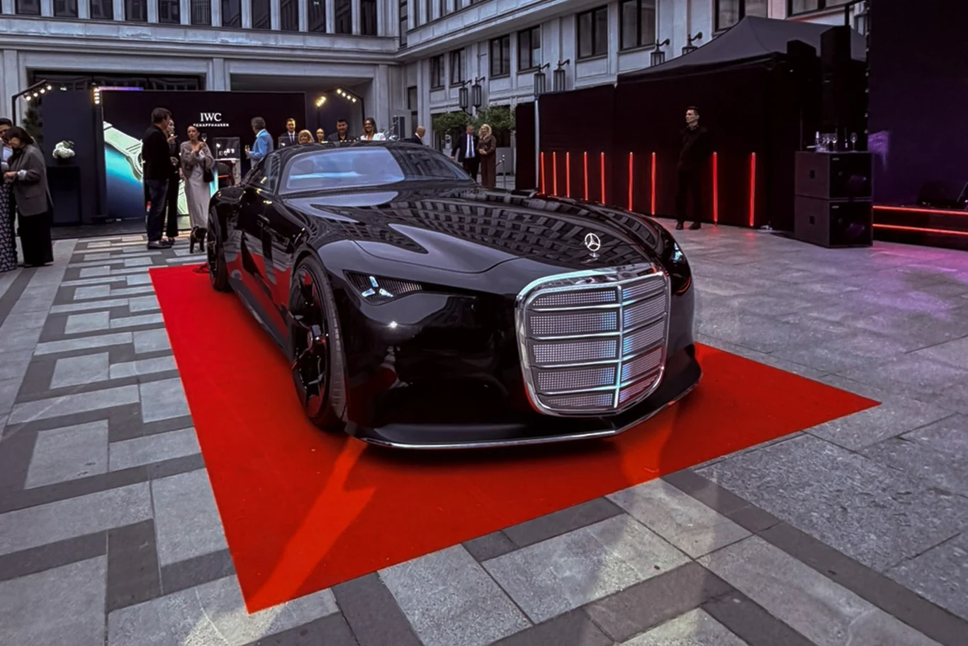 Mercedes-Benz Vision Iconic to tylko koncept, ale na żywo robi niesamowite wrażenie. Czarny, sportowy samochód Mercedes o futurystycznym designie ustawiony na czerwonym dywanie w otoczeniu nowoczesnych budynków i obserwujących osób.
