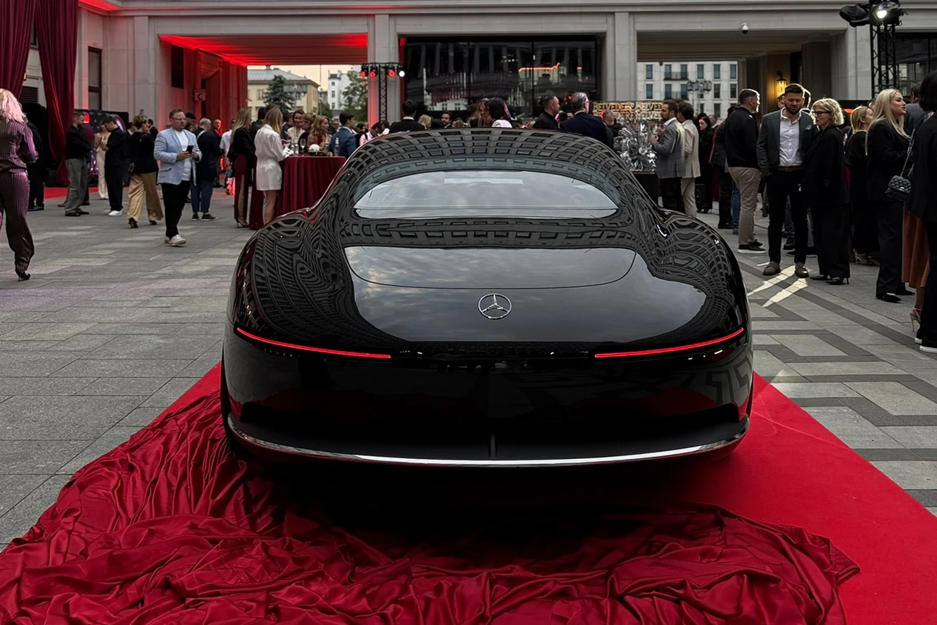 Mercedes-Benz Vision Iconic to pokaz możliwości niemieckiej marki. Klimat epoki art déco z lat 30. z high-techem. Luksusowy czarny samochód marki Mercedes zaprezentowany na czerwonym materiale, otoczony przez tłum elegancko ubranych osób podczas wydarzenia plenerowego.