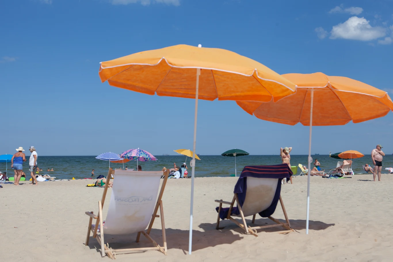 Dwie rozłożone pomarańczowe parasole, pod którymi stoją leżaki, na piaszczystej plaży z widokiem na morze oraz plażowiczami rozlokowanymi w tle.