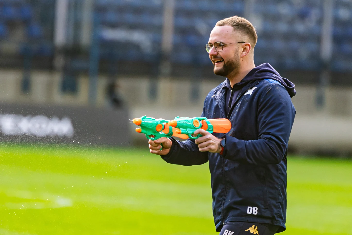 Młody mężczyzna w okularach i sportowej bluzie z uśmiechem trzyma dwie zabawkowe pistolety na wodę na zielonym boisku.