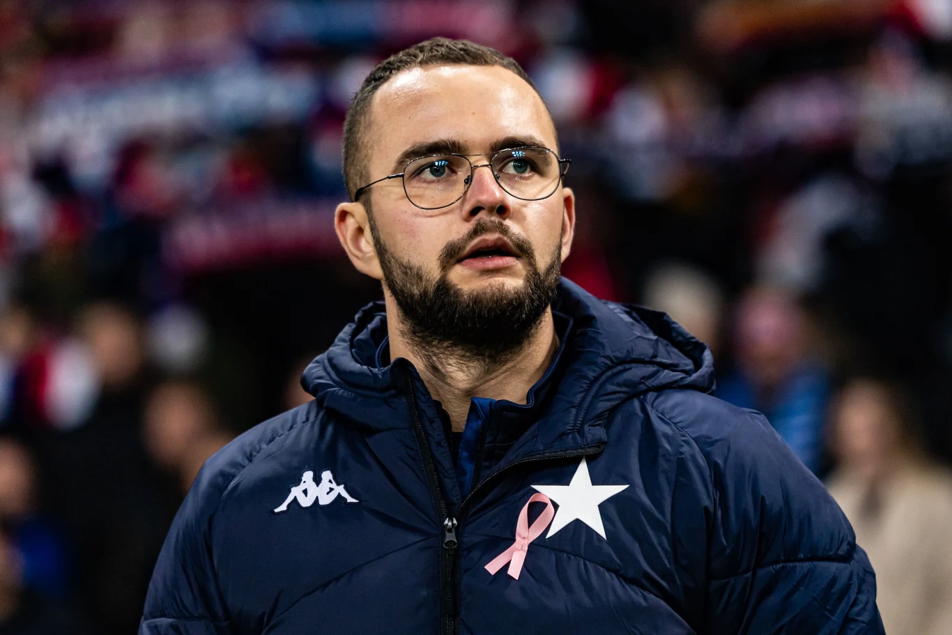 Mężczyzna w granatowej kurtce sportowej z białą gwiazdą i różową wstążką, w okularach, na tle rozmytego stadionu pełnego kibiców.