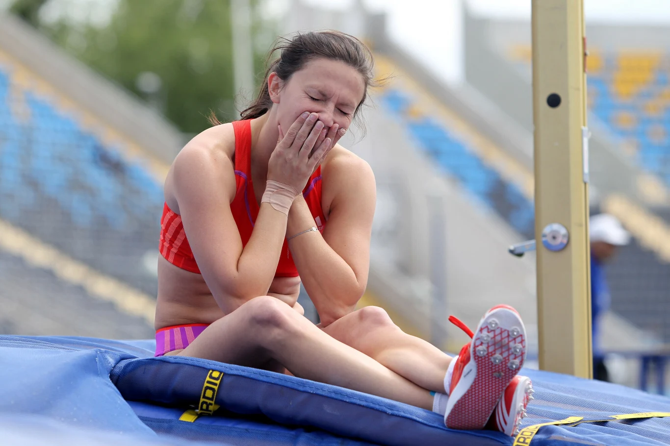 Zawodniczka siedzi na materacu lekkoatletycznym z dłońmi przy twarzy, wyrażając silne emocje i zmęczenie po skoku wzwyż.