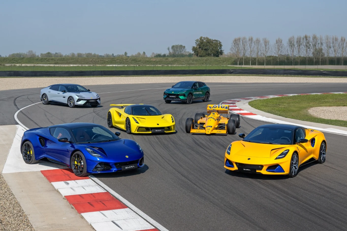 Pięć samochodów sportowych Lotus, w różnych kolorach, ustawionych obok siebie na torze wyścigowym, w tym jeden klasyczny bolid Formuły 1 i nowoczesne modele produkcyjne.
