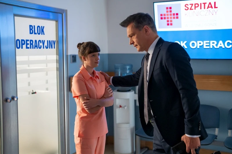 Czy Matylda zgodzi się przedstawić ukochanego ojcu? Dwójka osób rozmawia przed wejściem do bloku operacyjnego w szpitalu, mężczyzna w garniturze kładzie rękę na ramieniu kobiety ubranej w różowy strój medyczny, w tle widoczny jest ekran z nazwą szpitala.