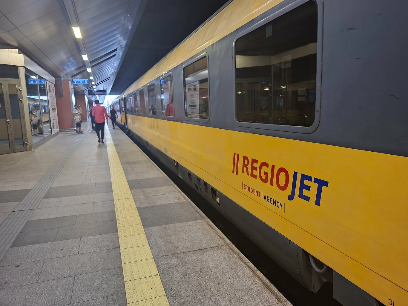 Jaskrawożółty pociąg RegioJet stoi na peronie dworca, przy którym idzie pojedynczy pasażer w czerwonej kurtce.