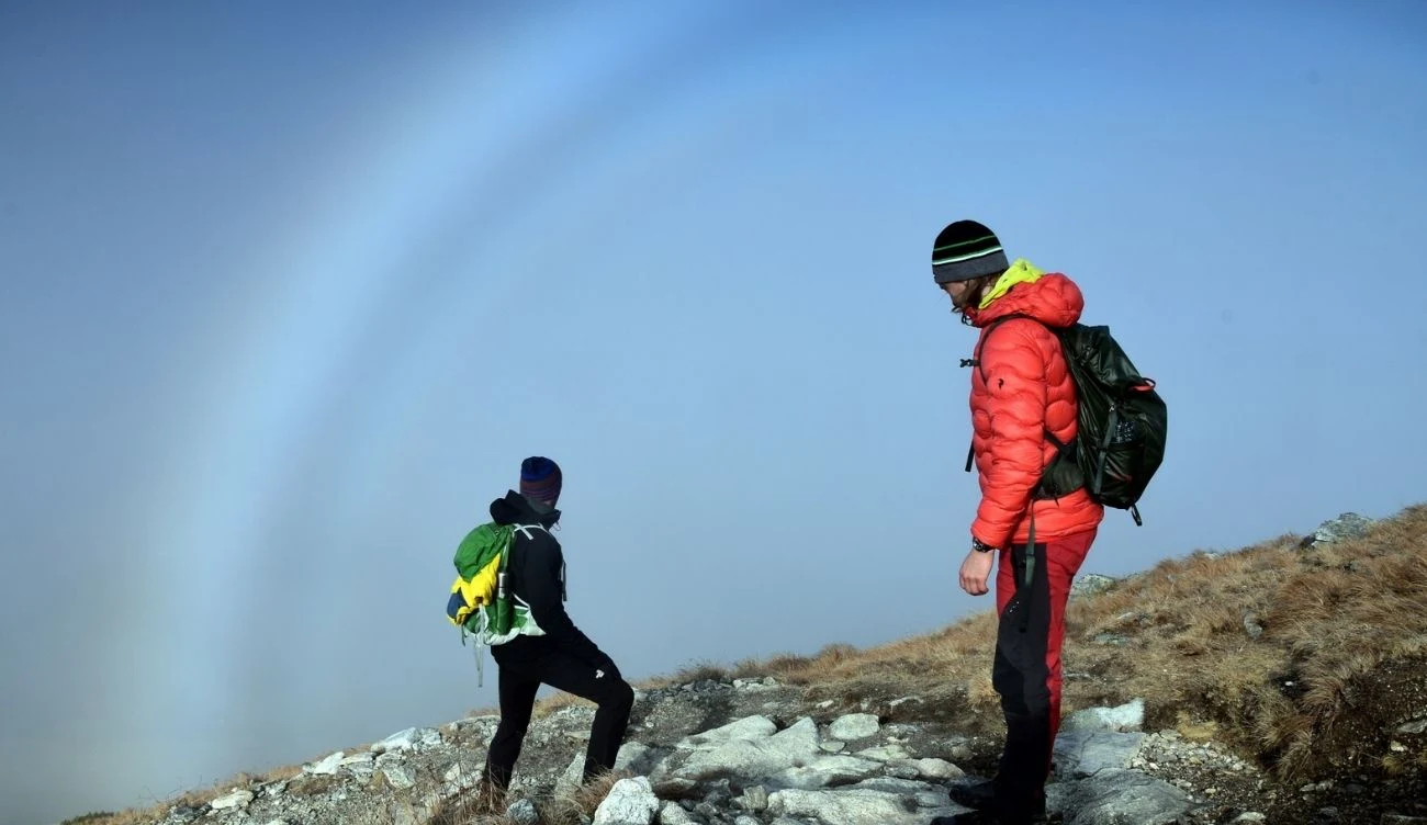 Fog bow, czyli biała tęcza, to wyjątkowo rzadkie zjawisko Dwóch turystów z plecakami na górskim szlaku; w tle biała tęcza na zamglonym niebie.