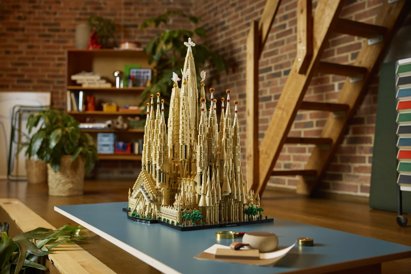 Model katedry Sagrada Familia zbudowany z klocków LEGO ustawiony na stole, w tle nowoczesne wnętrze z regałem i roślinami.