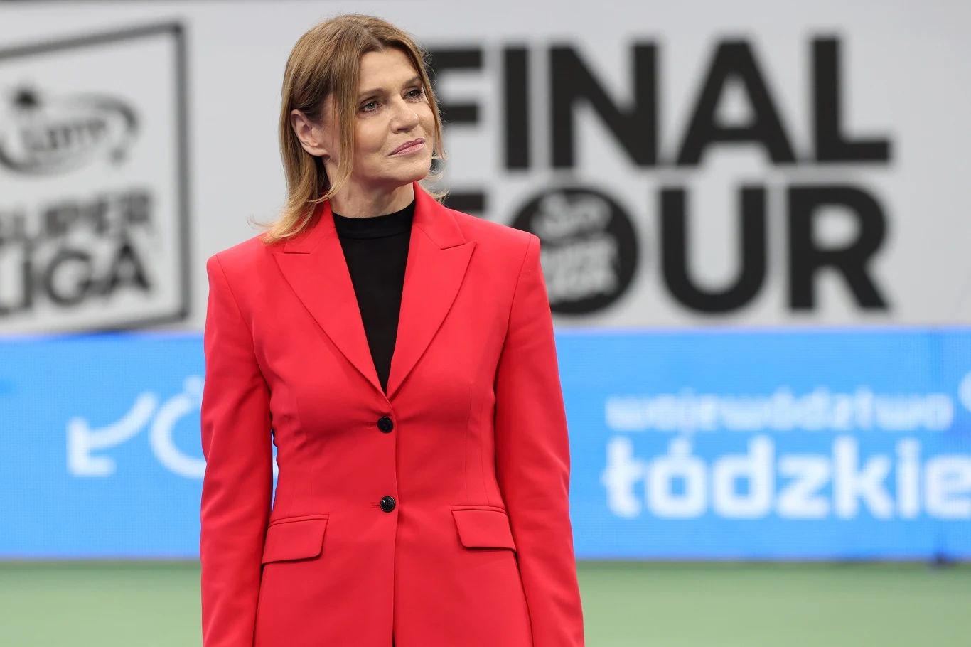 Kobieta w czerwonym garniturze stoi na tle banerów z napisem 'FINAL TOUR' oraz niebieskim wyświetlaczem z napisem 'łódzkie'.