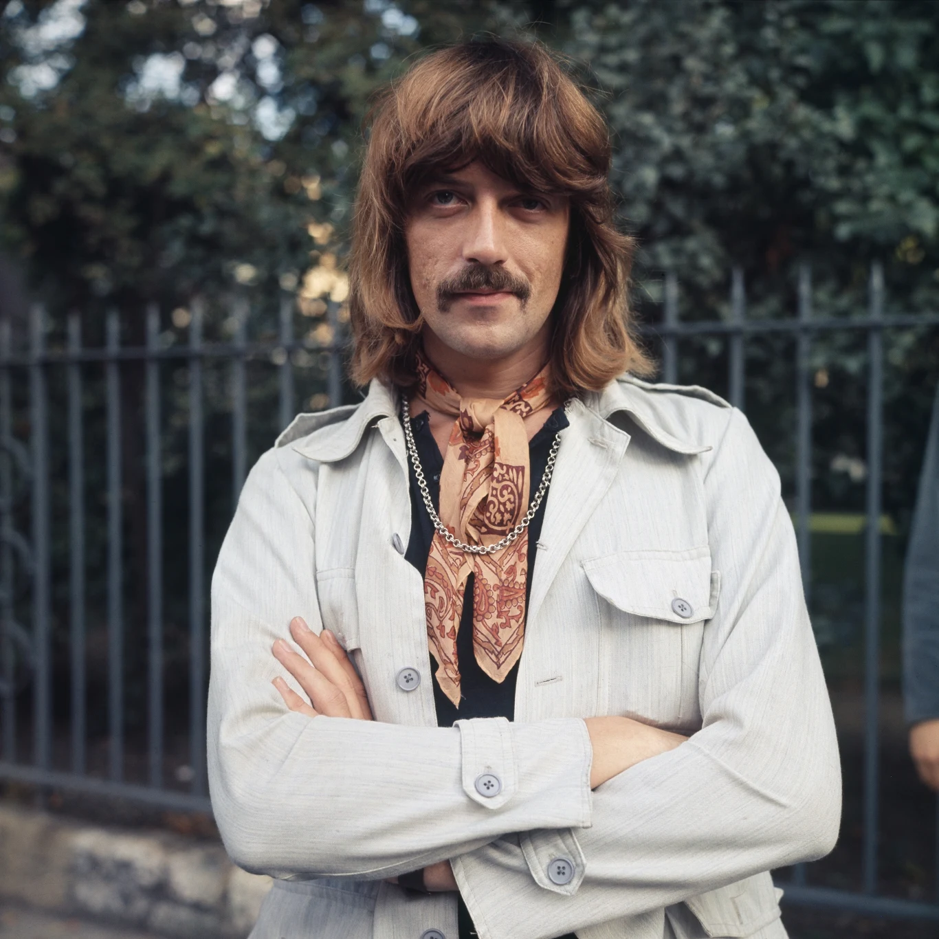Jon Lord (Deep Purple) w 1969 r. Mężczyzna z wąsami i długimi włosami w jasnej kurtce, z ramionami skrzyżowanymi na piersi, na szyi kolorowa apaszka i łańcuszek, w tle metalowe ogrodzenie i drzewa.