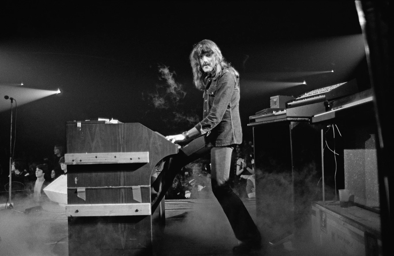 Jon Lord (Deep Purple) w 1974 r. Muzyk grający na klawiszach na scenie, w otoczeniu dymu scenicznego oraz oświetlenia koncertowego skierowanego w jego stronę.