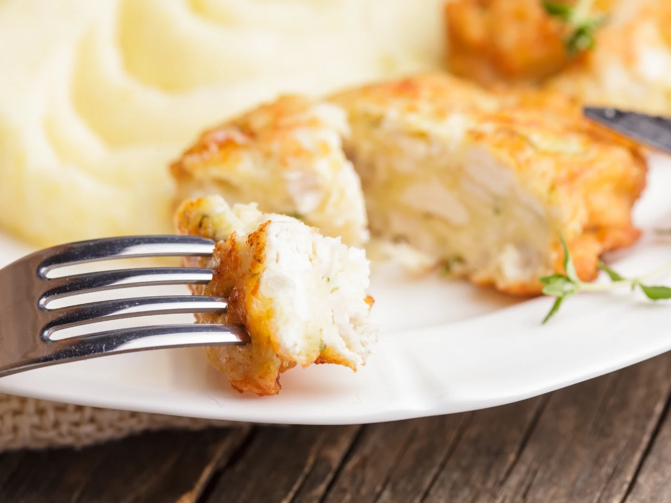 Cielęcina jak żadne inne mięso chroni serce i układ krwionośny Kawałek panierowanego mięsa na widelcu, z puree ziemniaczanym i ziołami na białym talerzu.