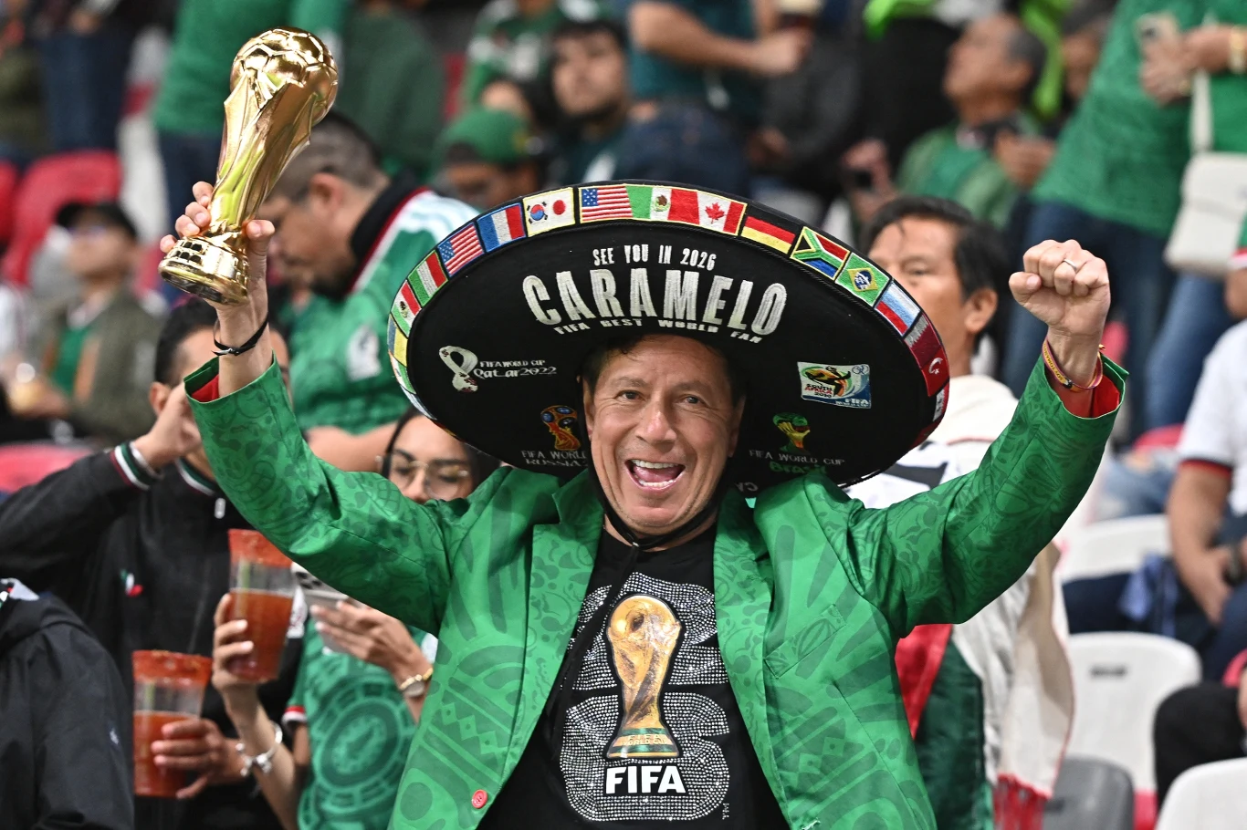 Kibic reprezentacji Meksyku z symbolami mundialu 2026 Uśmiechnięty mężczyzna w zielonej marynarce i dużym, ozdobionym logotypami sombrero unosi w górę ręce i trzyma replikę Pucharu Świata FIFA, otoczony kibicami na stadionie.