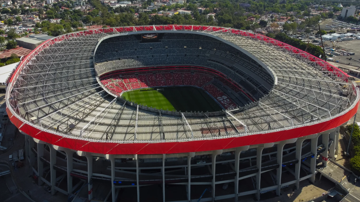Nowoczesny stadion piłkarski o owalnym kształcie, z zadaszeniem nad trybunami i zieloną murawą pośrodku, wypełniony kibicami na widowni.