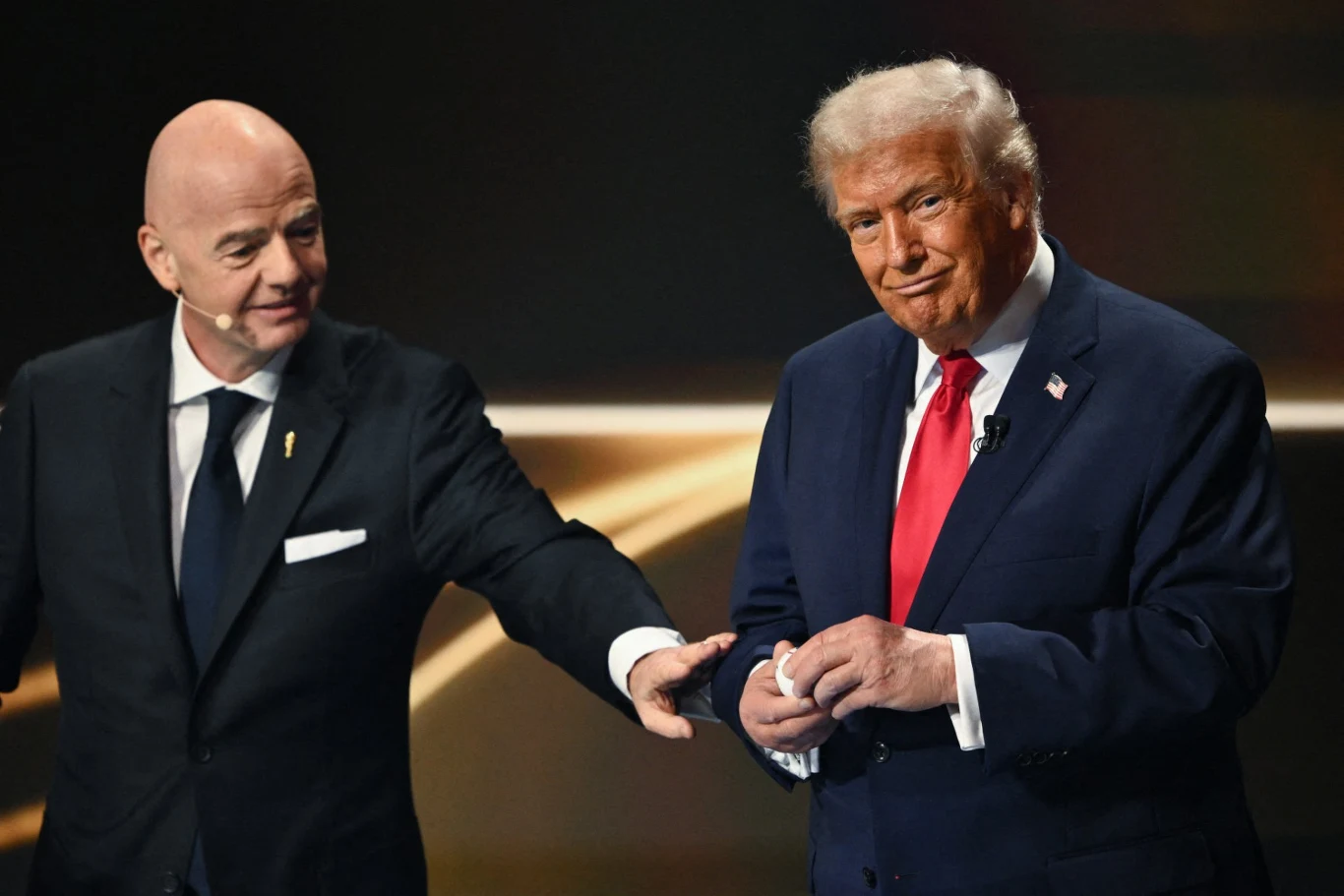 Dwóch elegancko ubranych mężczyzn w garniturach, jeden wskazuje drugiego w kierunku sceny, drugi stoi z zaciśniętymi dłońmi i lekko uśmiechniętą miną.