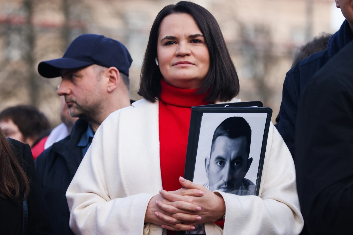 Liderka białoruskiej opozycji Swiatłana Cichanouska Swiatłana Cichanouska w białym płaszczu i czerwonym golfie trzyma czarno-białe zdjęcie mężczyzny na tle miasta.