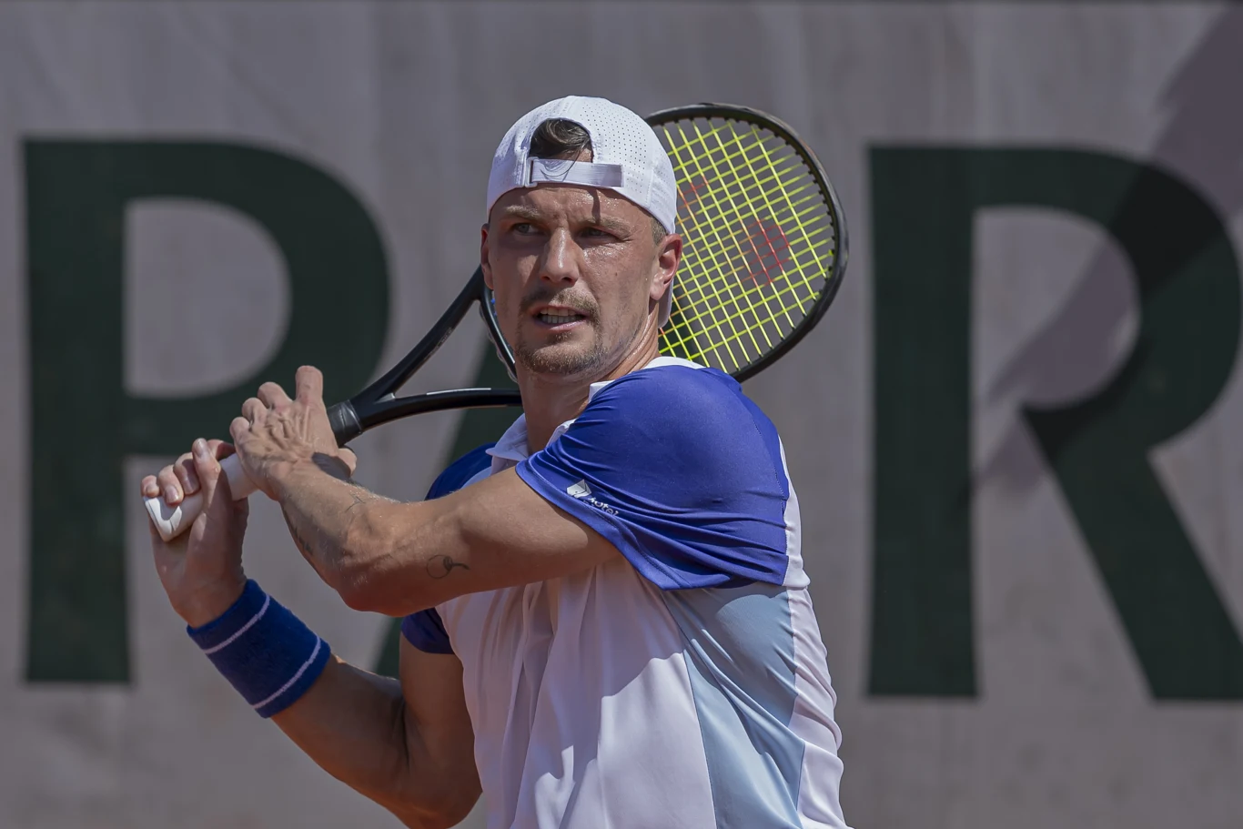 tenisista w białej czapce i sportowej koszulce w niebiesko-białych barwach przygotowuje się do uderzenia rakietą podczas meczu na korcie ziemnym