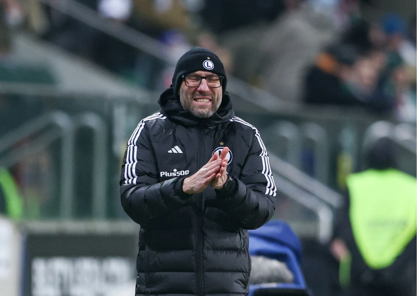 Uśmiechnięty trener w czarnej kurtce i czapce z logo sportowego, stojący przy linii bocznej boiska na tle stadionu oraz rozmytych postaci w tle.