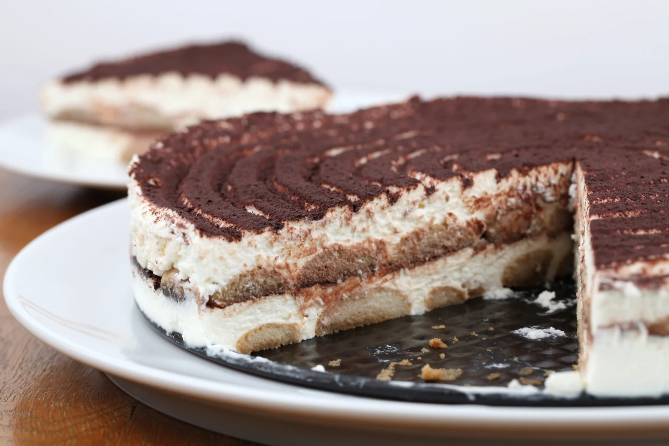 Szybkie ciasto tiramisu nie wymaga pieczenia, łączy w sobie krem na serku mascarpone oraz biszkopty nasączone w kawie Okrągłe ciasto tiramisu bez pieczenia, przekrojone, widoczne warstwy biszkoptów, mascarpone i kakao na talerzu.