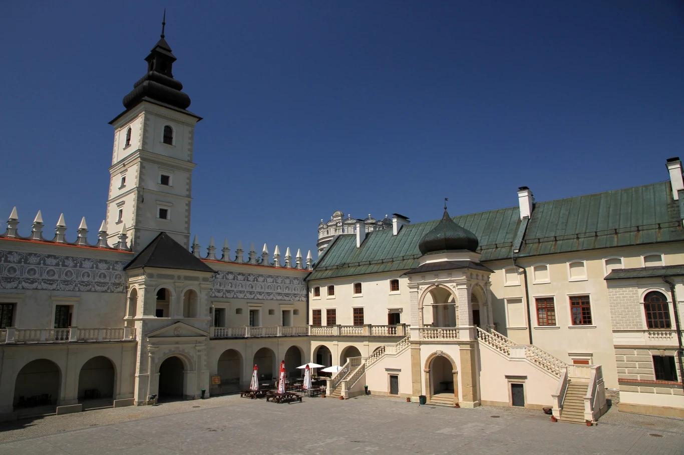 Dziedziniec renesansowego zamku z wysoką wieżą, arkadowymi krużgankami i schodami prowadzącymi do głównego wejścia, na placu kilka rozstawionych parasoli i stolików.