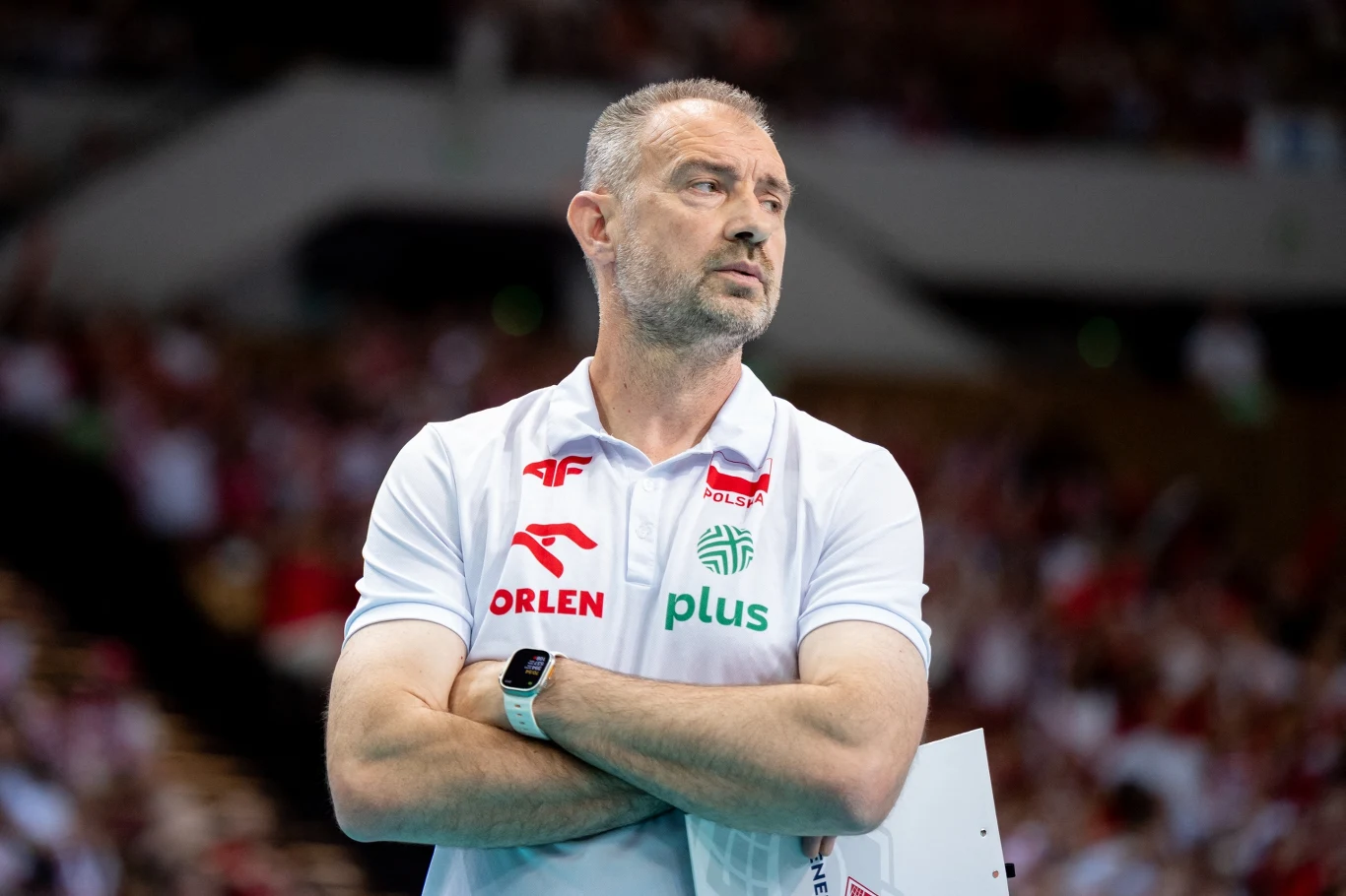 Trener drużyny siatkarskiej w białej koszulce z logo sponsorów, z założonymi rękami i skupionym wyrazem twarzy, znajduje się na hali sportowej podczas meczu.