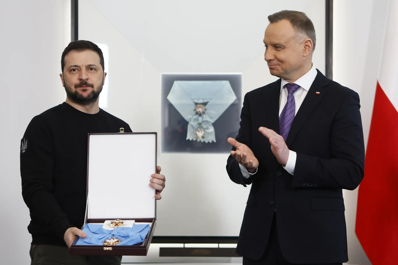Mężczyzna w ciemnym swetrze trzyma otwartą kasetkę z odznaczeniem, obok stoi drugi mężczyzna w garniturze i fioletowym krawacie, który klaszcze; w tle widoczne polskie barwy narodowe i powiększone zdjęcie tego samego odznaczenia.