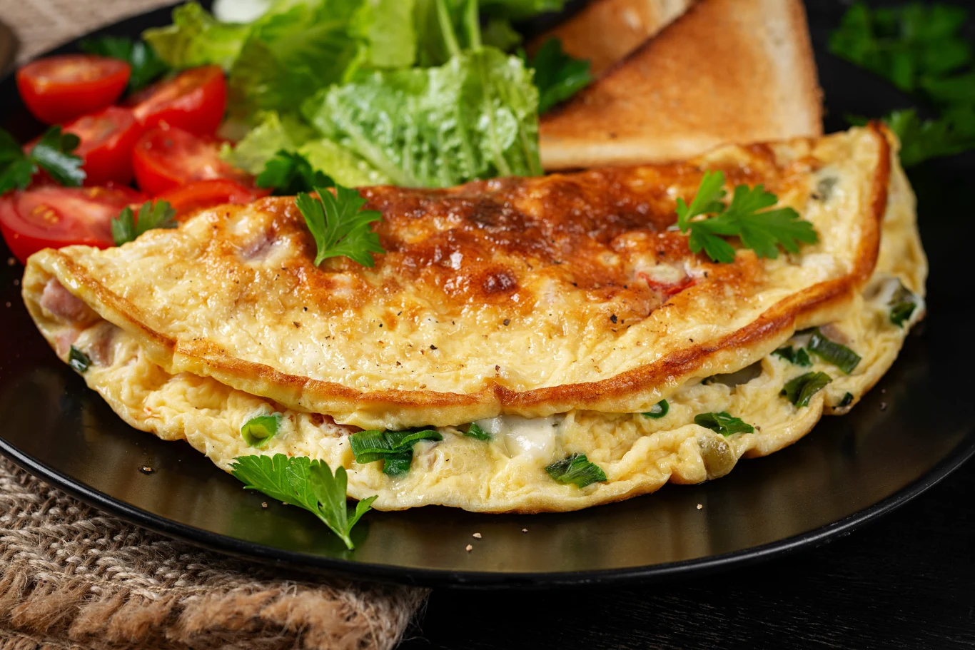 Omlet z ciecierzycą nasyci na długo nawet bez chleba. Omlet z ciecierzycą i ziołami na czarnym talerzu, obok sałata, plasterki pomidora i kromki grzanki.