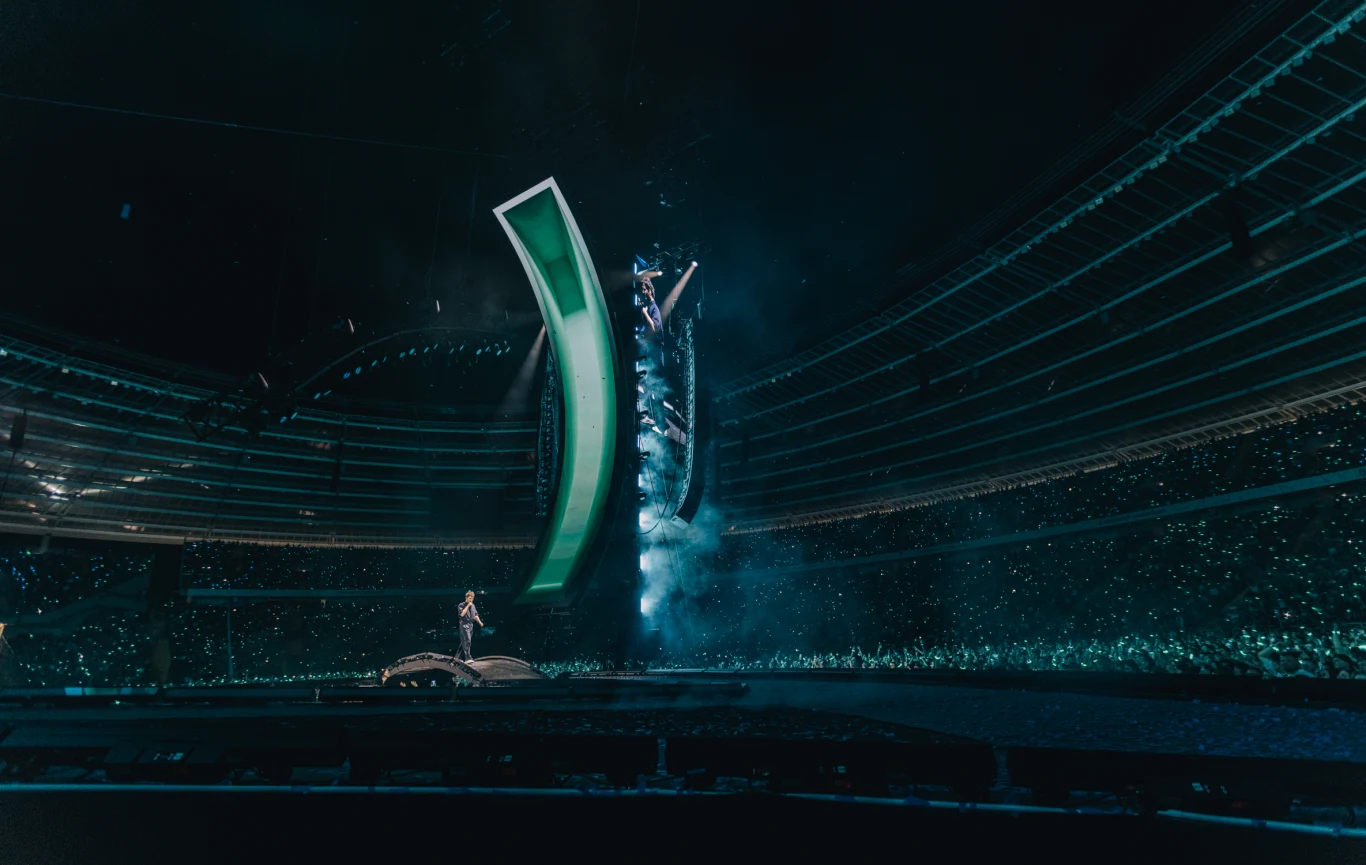 Ogromna scena koncertowa w nowoczesnym stadionie, oświetlona efektownymi światłami, na której występują artyści przed tłumem rozświetlonym tysiącami drobnych światełek.