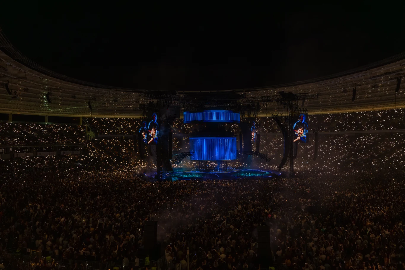 Tłum ludzi zgromadzonych na stadionie podczas nocnego koncertu, scena podświetlona na niebiesko, duże ekrany wyświetlają artystę, a tysiące światełek tworzą wyjątkową atmosferę.