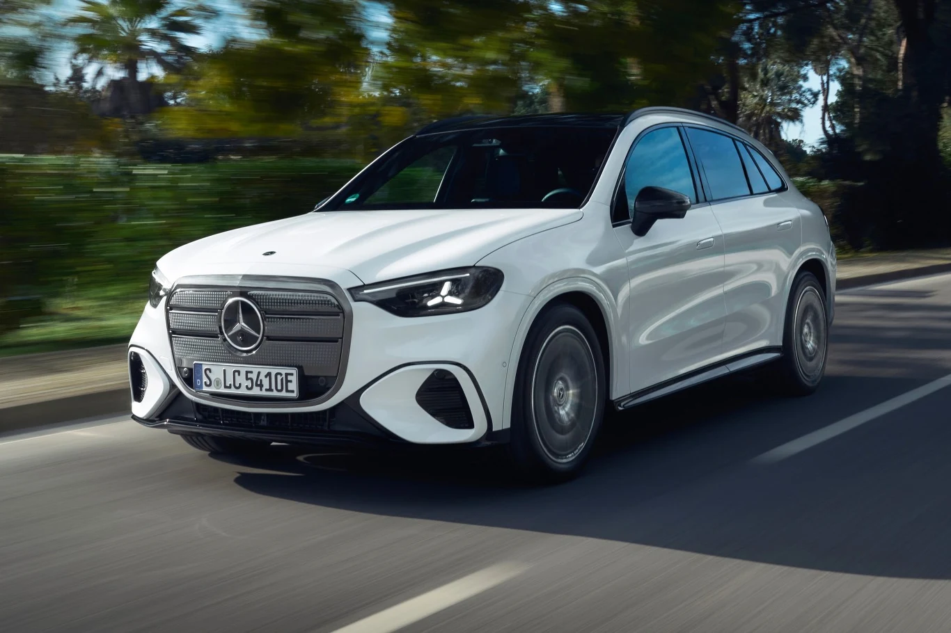 Nowoczesny biały samochód elektryczny marki Mercedes-Benz jedzie po dwupasmowej, asfaltowej drodze otoczonej zielenią.