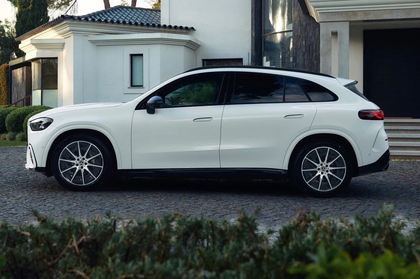 Biały SUV o nowoczesnej sylwetce zaparkowany na brukowanym podjeździe przed eleganckim, jasnym domem z wyrazistą architekturą.