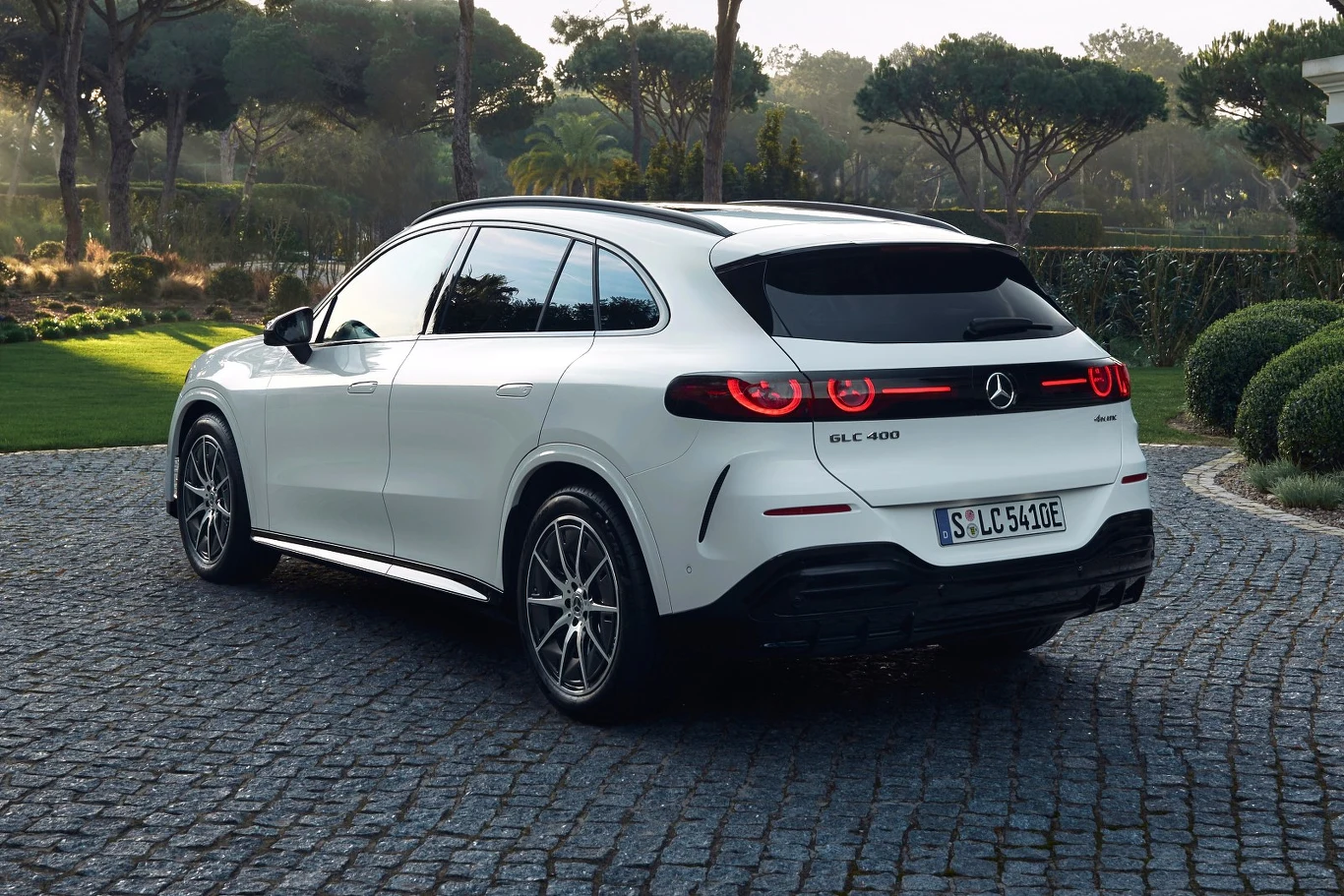 Biały SUV marki Mercedes-Benz GLC 400e stoi na brukowanej nawierzchni, otoczony zadbaną zielenią i drzewami w tle.