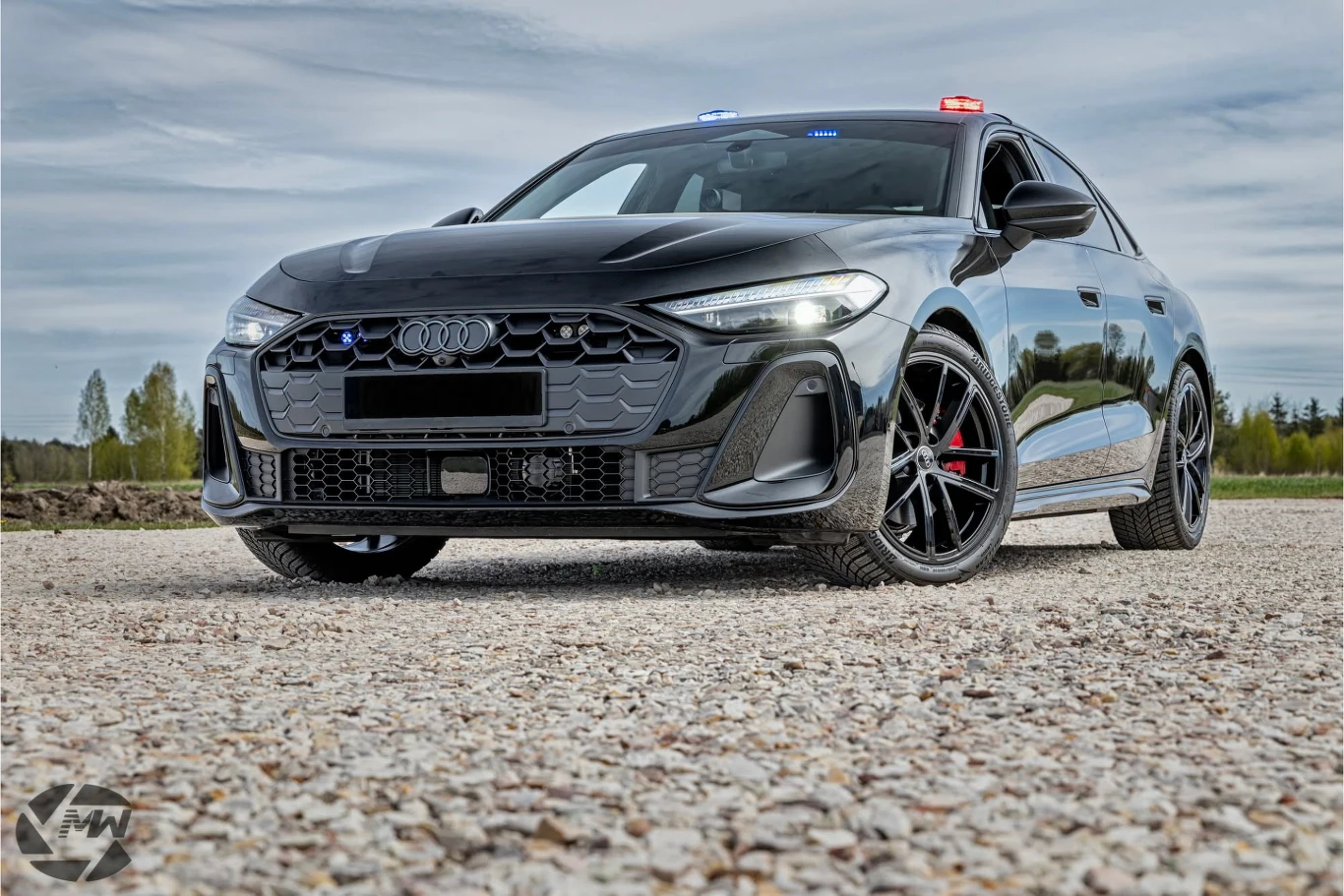 Audi A5 w konfiguracji nieoznakowanego radiowozu. Czarne Audi A5 z policyjnym oznakowaniem świetlnym na dachu, sfotografowane z przodu na żwirowej nawierzchni.