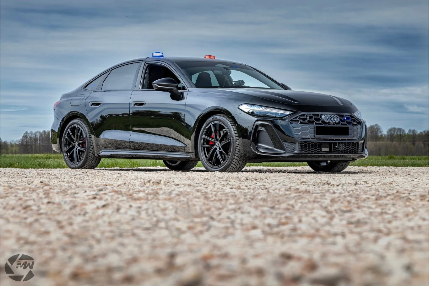 Prezentowane Audi A5 jest na razie wyłącznie propozycją - spełnia jednak wszystkie wymagania policji. Czarny sportowy samochód marki Audi z niebieskim i czerwonym sygnałem świetlnym na dachu, stojący na otwartej przestrzeni z widokiem na pole i drzewa w oddali.