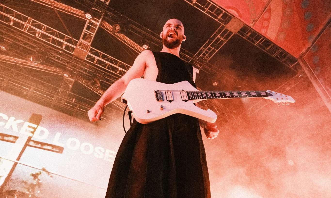 Mężczyzna w czarnej stylizacji trzymający białą gitarę elektryczną na scenie koncertowej, oświetlony scenicznymi światłami i otoczony dymem.