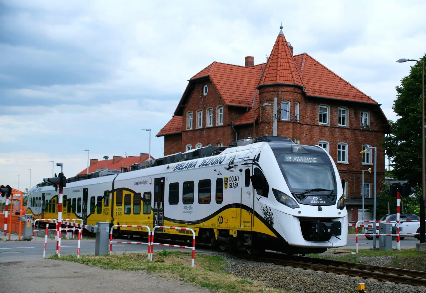 Pierwszy przejazd w Góry Sowie. Pociągi Kolei Dolnośląskich od niedzieli. Pociąg Kolei Dolnośląskich przejeżdża przez przejazd kolejowy obok ceglanego budynku ze spadzistym dachem.