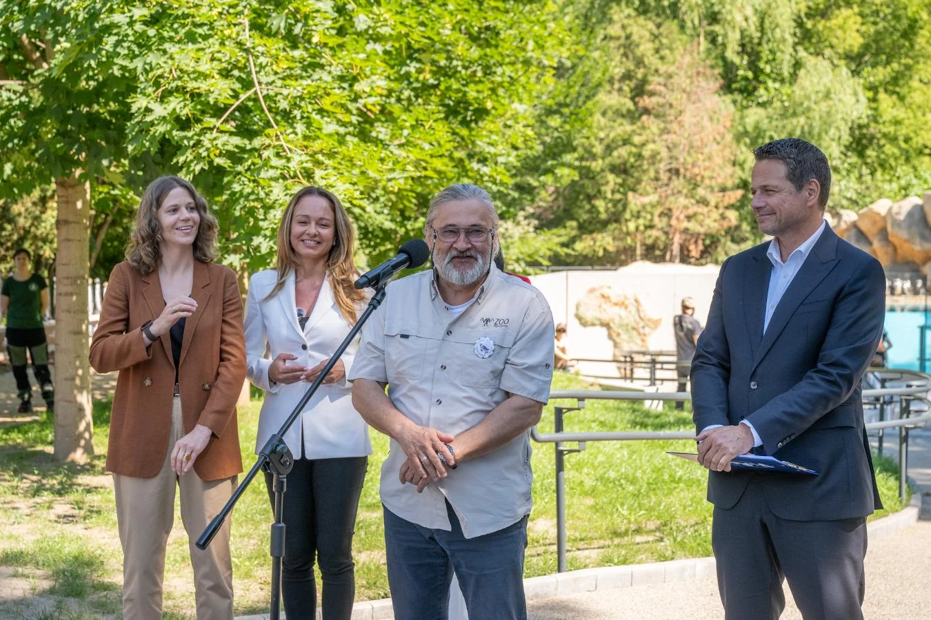 Dyrektor warszawskiego ogrodu zoologicznego dr Andrzej Kruszewicz oraz prezydent Warszawy Rafał Trzaskowski podczas otwarcia nowego wybiegu dla pingwinów Cztery osoby stoją na świeżym powietrzu przed mikrofonem, dwie kobiety i dwóch mężczyzn uśmiechają się, otoczeni zielenią drzew i słonecznym światłem.