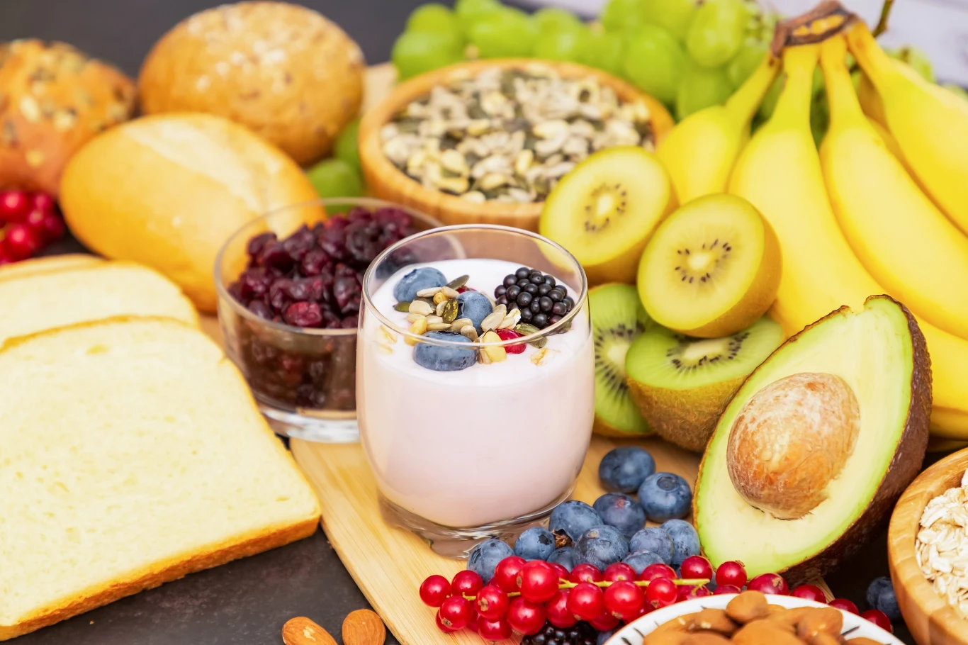 Jogurt z owocami i orzechami otoczony plasterkami awokado, kiwi, bananami, winogronami, pieczywem, bułkami, jagodami oraz pestkami dyni na drewnianej desce.