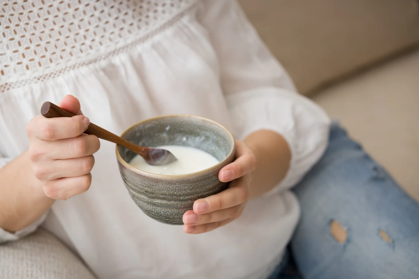 Osoba trzymająca ceramiczną miseczkę z jogurtem naturalnym i łyżeczką, ubrana w białą bluzkę z ażurowym wzorem i niebieskie dżinsy z przetarciami.