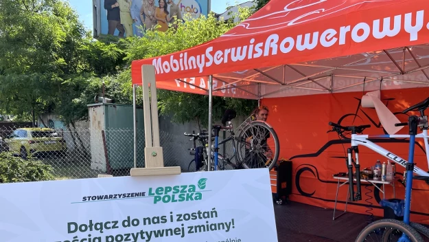 Stoisko mobilnego serwisu rowerowego z charakterystycznym czerwonym namiotem, tablicą informacyjną zachęcającą do dbania o środowisko oraz serwisowanymi rowerami na stojakach.