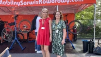 Dwie kobiety stoją przed stoiskiem mobilnego serwisu rowerowego, w tle widoczne podniesione na stojakach rowery oraz mechanik pracujący przy jednym z nich.