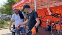 Dwóch mężczyzn przy stoisku serwisowym omawia szczegóły konserwacji roweru, jeden z nich wskazuje elementy przy kierownicy, na drugim planie widoczne narzędzia oraz części rowerowe.