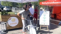 Kobieta w jasnych ubraniach pedałuje na rowerze stacjonarnym ustawionym pod namiotem, obok stoi blender i tablica informacyjna, w tle widoczne logo polsat oraz banner Mobilny Serwis Rowerowy.
