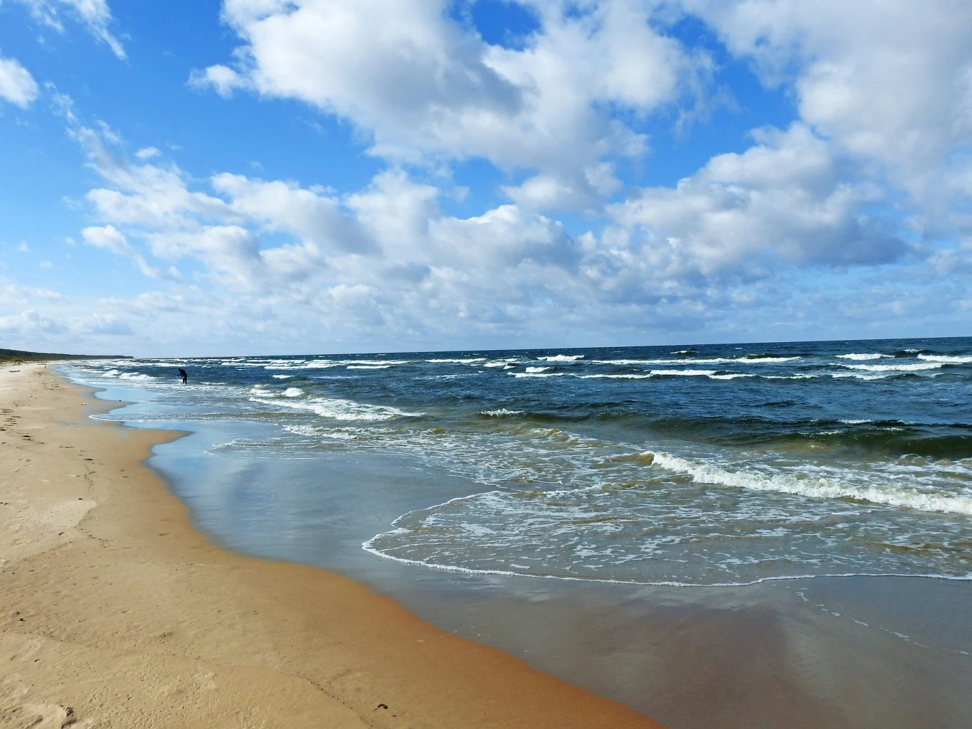 Spokojne morze może okazać się bardzo niebezpieczne Piaszczysta plaża nad morzem z delikatnymi falami i częściowo zachmurzonym, błękitnym niebem.