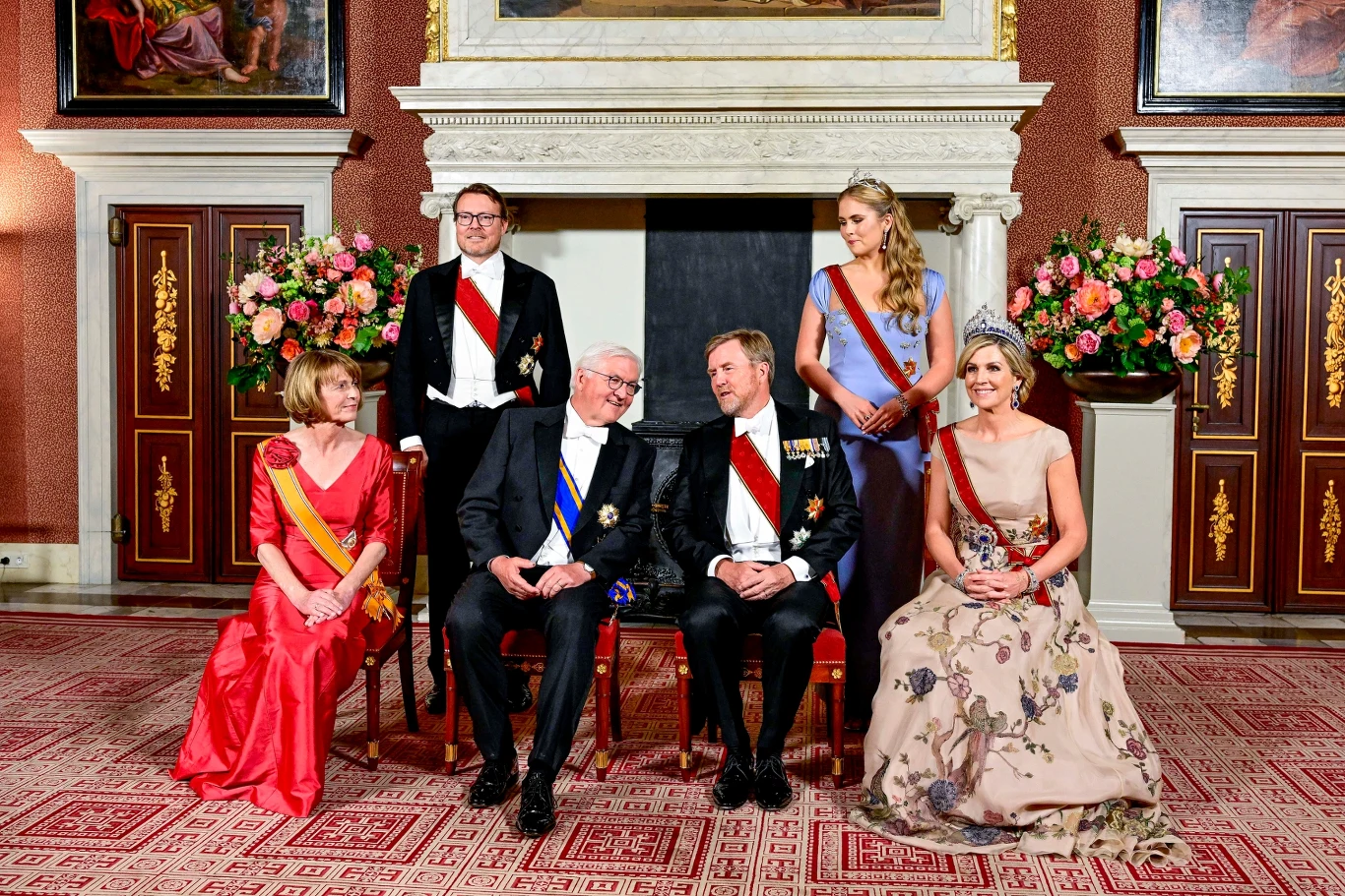 Sześć elegancko ubranych osób siedzi i stoi przed kominkiem, otoczonych bukietami kwiatów; większość nosi odznaczenia i szarfy świadczące o oficjalnym charakterze spotkania.