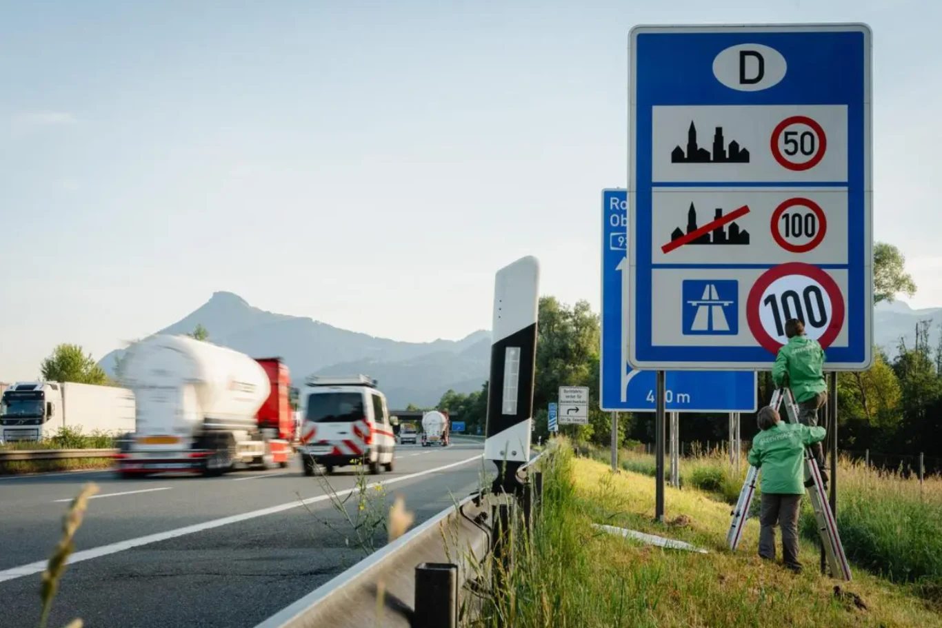Nowy limit prędkości na niemieckich autostradach nie jest efektem zmian przepisów. Dwie osoby zmieniające oznaczenie ograniczenia prędkości na dużym znaku drogowym przy autostradzie, obok szybkie samochody i ciężarówki na tle górskiego krajobrazu.