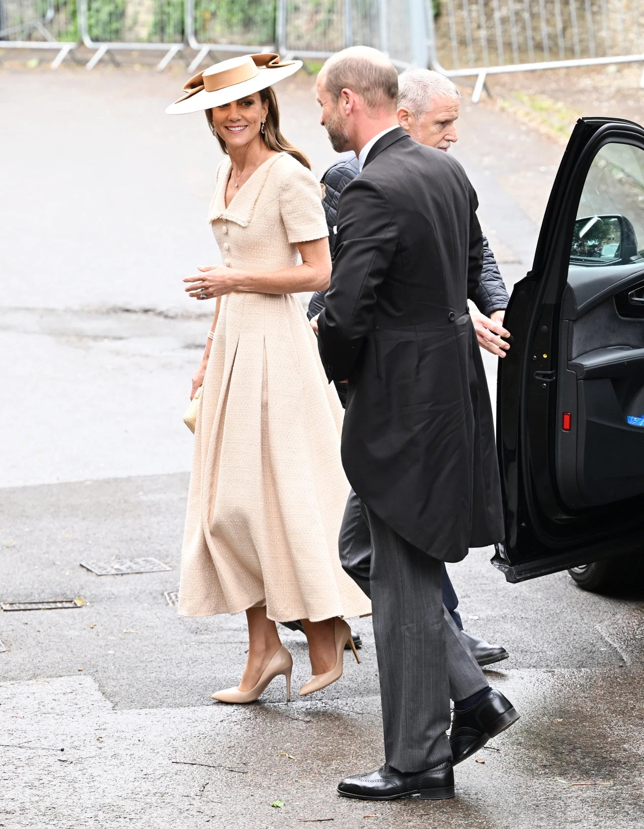 Elegancko ubrana kobieta w beżowej sukience i kapeluszu stoi na chodniku obok mężczyzny w czarnym fraku, obok otwartych drzwi samochodu.