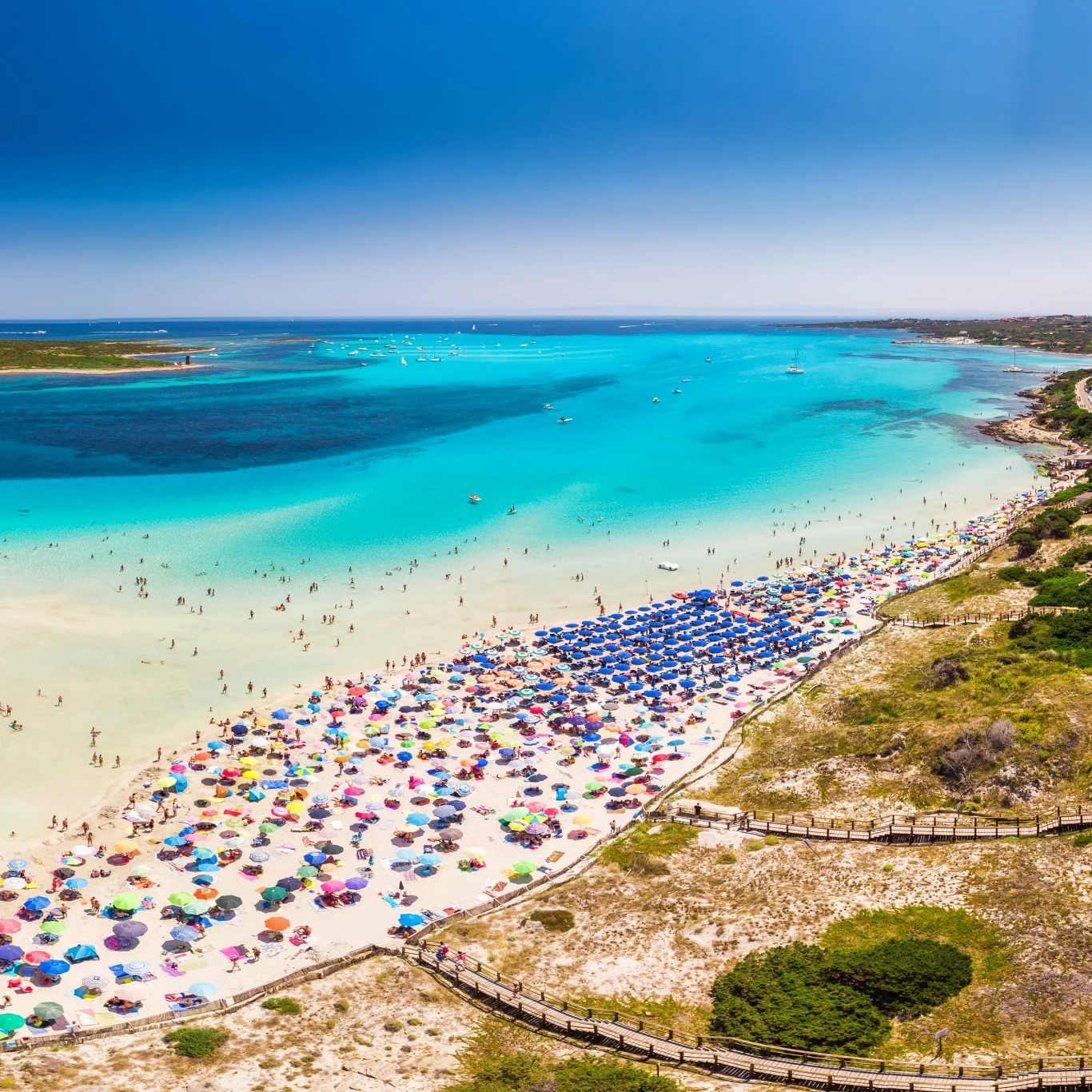 Tłum ludzi korzystających z kolorowych parasoli na szerokiej, piaszczystej plaży przy przejrzystym, turkusowym morzu.